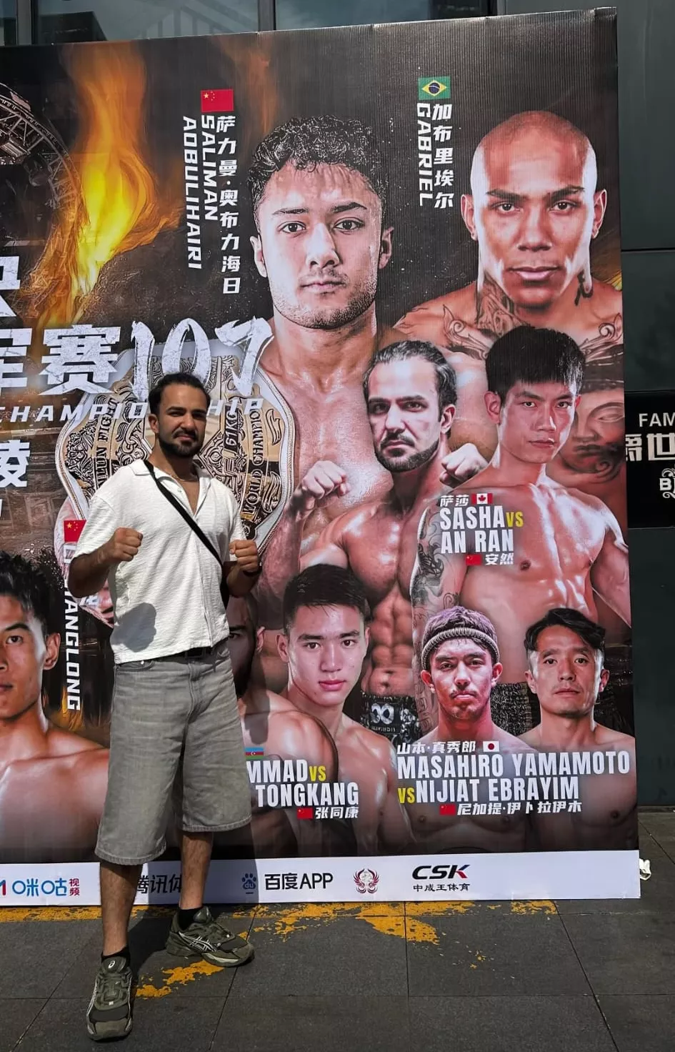 Sasha Tadayoni standing in front of the official Kunlun Fight 107 poster showing Sasha of Canada vs An Ran